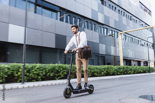 bearded businessman in formal wear riding electric scooter and holding bag near building