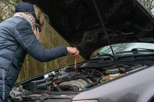 Young blonde woman in a winter down jacket in a yellow vest, talking on the phone and checking the engine oil level of the car. Photo from the back