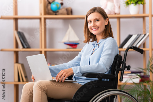 Happy female psychologist with laptop looking at camera while sitting in wheelchair on blurred background