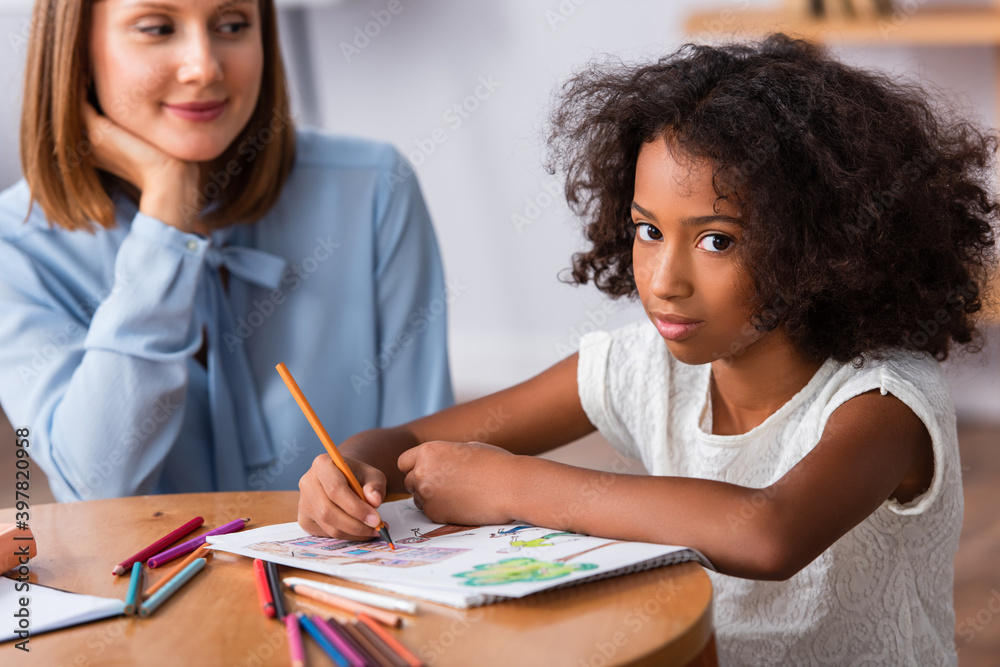 African american girl looking at camera while drawing with colored pencils near positive psychologist on blurred background African american girl looking at camera while drawing with colored pencils near positive psychologist on blurred background