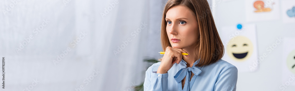 Thoughtful female psychologist holding pen and looking away with blurred office on background, banner Thoughtful female psychologist holding pen and looking away with blurred office on background, banner