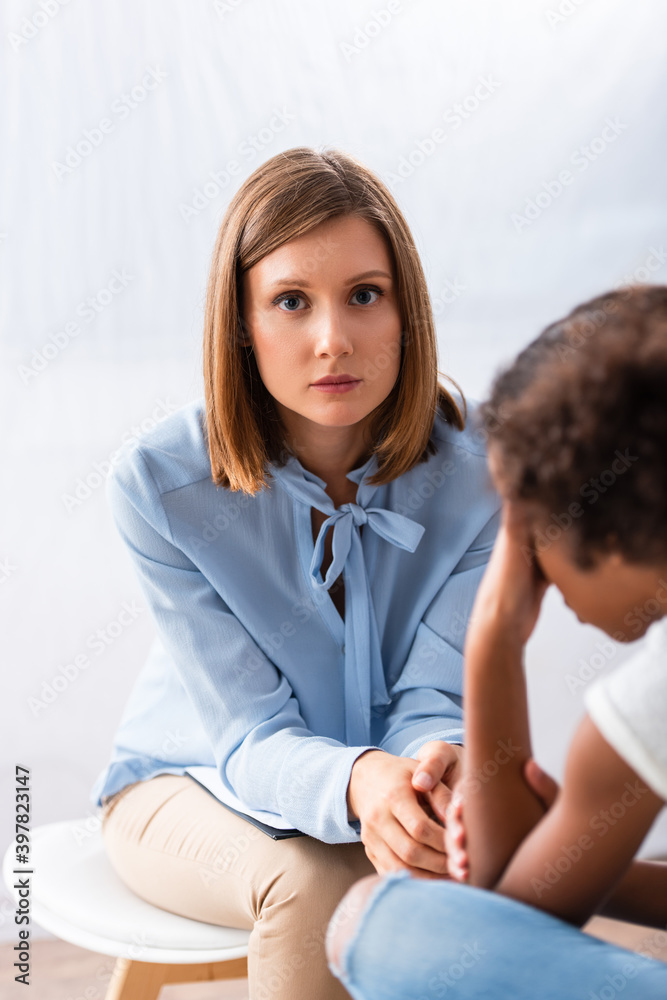 Female psychologist looking at camera while sitting on chair near blurred african american girl crying on foreground Female psychologist looking at camera while sitting on chair near blurred african american girl crying on foreground