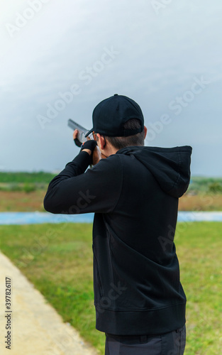 A man in dark clothes and a cap is aiming with a double-barreled gun. Back view. Skeet shooting.