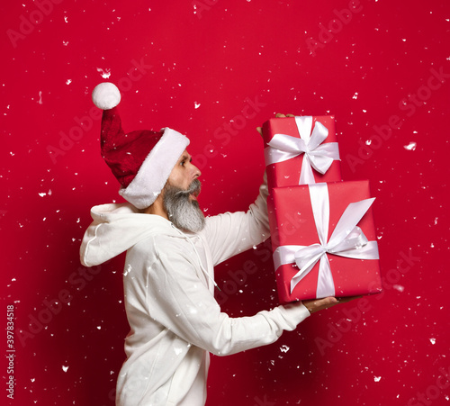 Cristmas presents. Side view of a senior bearded man hurrying to give gifts with a surprised expression on his face, against a red background. Gifts for the family. Surprise concept. snowy photo