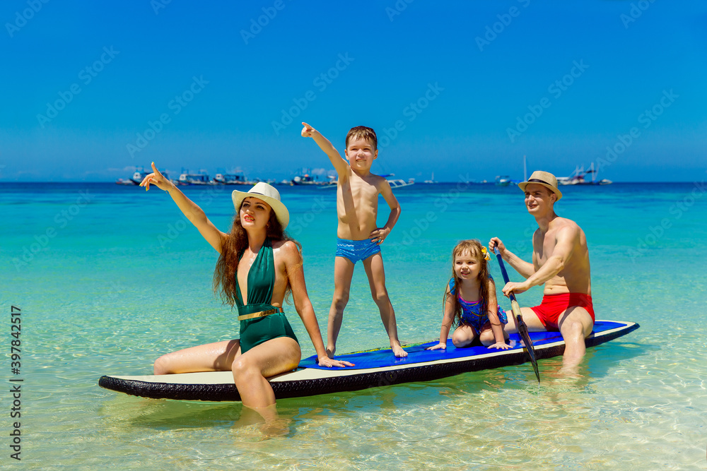 Obraz premium Happy family-mom, dad, little girl and boy have fun on a paddleboard in the tropical sea. The concept of travel and family holidays.