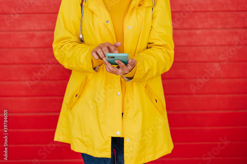 Woman in yellow raincoat using smartphone in front of red wall photo