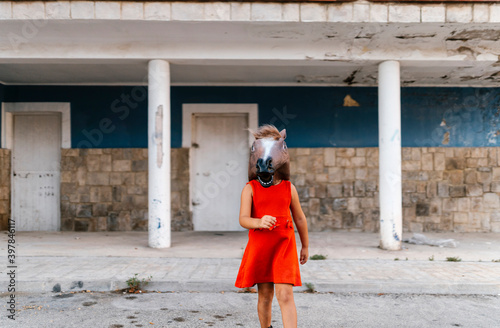 Little girl with a horse's head and a red dress walking away from an abandoned house photo