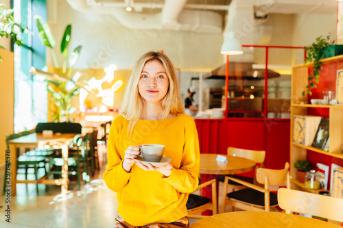 Portrait of blond caucasian female holding cup with hot caffeine beverage resting in cafe loft modern interior with green plants, funny hipster girl looking at camera enjoying mocha tea