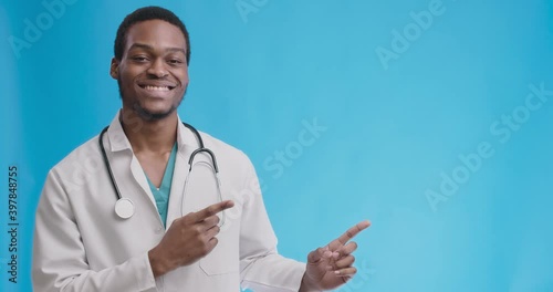 Cheerful african american professional doctor showing empty space for mock up, pointing aside aon blue studio background photo