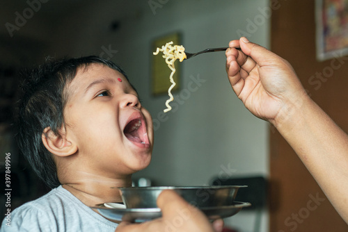 Portrait of a cute little Indian girl  2-3 years  while eating breakfast. She is seen opening her mouth and looking at ramen noodle fork while being spoon fed.