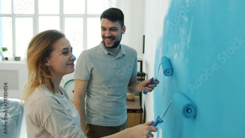 Playful guy is having fun with girlfriend touching her nose with dirty finger painting wall together in apartment. Renovation and relationship concept.