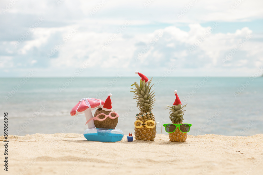 funny attractive pineapples and coconut in stylish sunglasses on the sand against turquoise sea. Wearing christmas hats. 