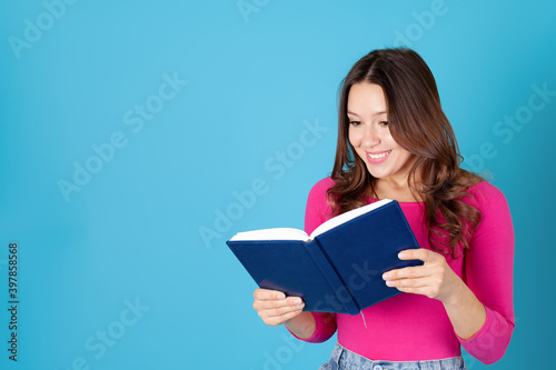 close-up portrait of a smiling young woman holding a diary or book in her hands and reading it with pleasure, isolated on a blue background.