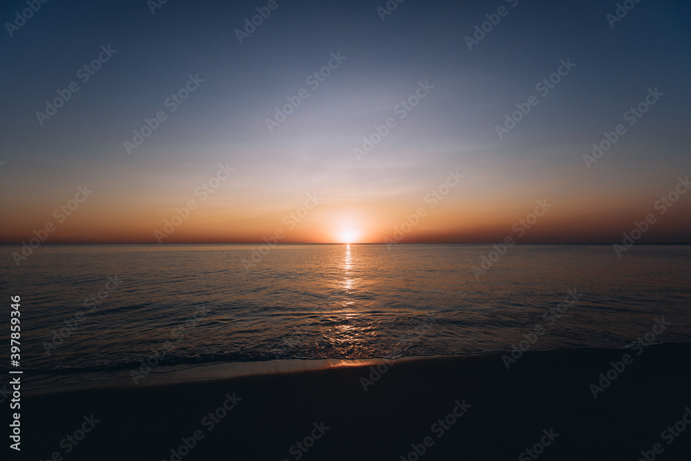Obraz premium Breathtaking summer sunset view on the beach. Wonderful sunset landscape at the deep dark sea and orange sky above it and calm waves are flowing on it.