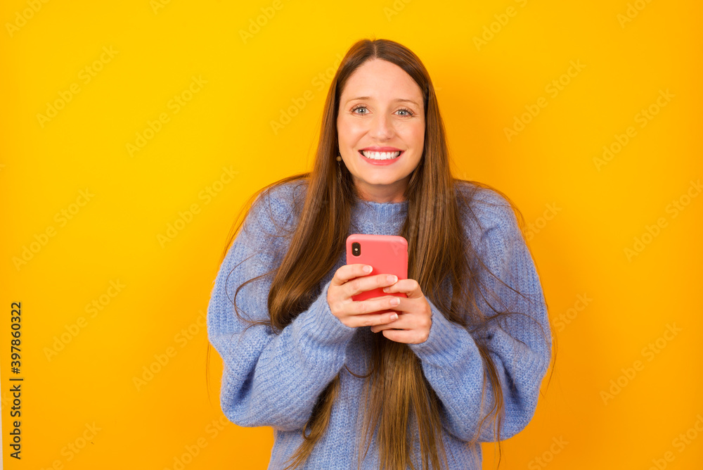 Φωτογραφία Excited Young beautiful Caucasian woman wearing blue sweater against yellow wall holding smartphone and looking amazed to the camera after receiving good news