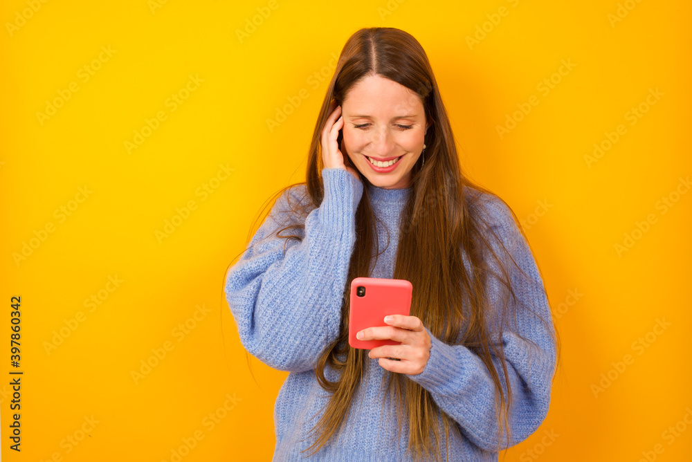 Φωτογραφία Happy Young beautiful Caucasian woman wearing blue sweater against yellow wall feels good while focused in screen of smartphone