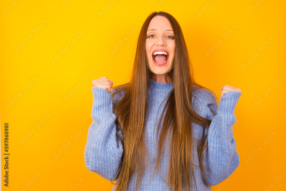 Young beautiful Caucasian woman wearing blue sweater against yellow wall celebrating surprised and amazed for success with arms raised and open eyes. Winner concept. Young beautiful Caucasian woman wearing blue sweater against yellow wall celebrating surprised and amazed for success with arms raised and open eyes. Winner concept.