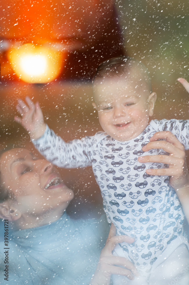 Obraz premium Happy mother and her baby boy looking throw the window and playing in warm home on winter day. Christmas time.