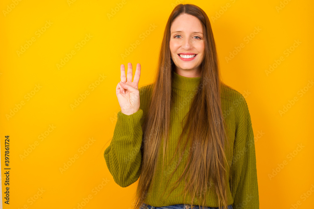Young beautiful Caucasian woman wearing green sweater against yellow wall showing and pointing up with fingers number three while smiling confident and happy.