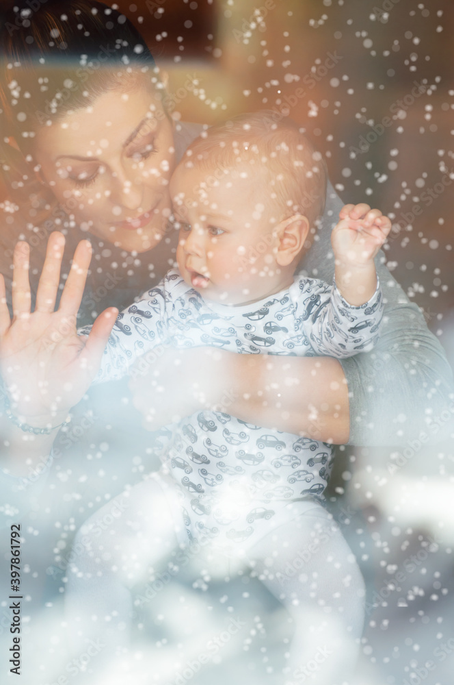 Happy mother and her baby boy looking throw the window and playing in warm home on winter day. Christmas time. Happy mother and her baby boy looking throw the window and playing in warm home on winter day. Christmas time.