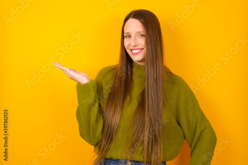 Young beautiful Caucasian woman wearing green sweater against yellow wall smiling cheerful presenting and pointing with palm of hand looking at the camera.