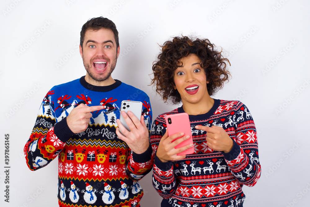 Young couple wearing Christmas sweater standing against white wall holding in hands showing new cell,