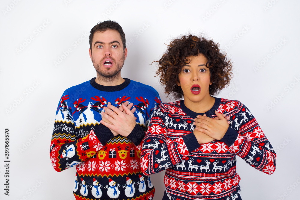 Scared Young couple wearing Christmas sweater looks with frightened expression, keeps hands on chest, being puzzled to notice something strange, People, hush reaction and emotions.