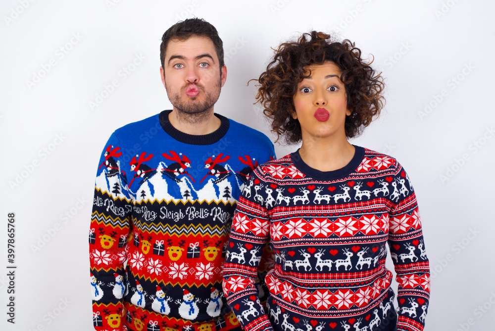 Shot of pleasant looking Young couple wearing Christmas sweater standing against white wall, pouts lips, looks at camera, Human facial expressions