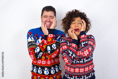 Doleful desperate crying Young couple wearing Christmas sweater standing against white wall, looks stressfully, frowns face, feels lonely and anxious