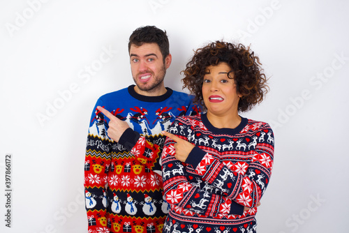 Young couple wearing Christmas sweater standing against white wall Pointing aside worried and nervous with forefinger, concern and surprise concept.