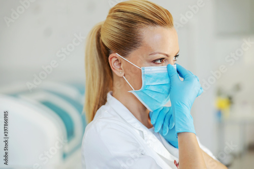 Portrait of attractive blond lab assistant with rubber gloves standing in hospital and putting sterile face mask on.