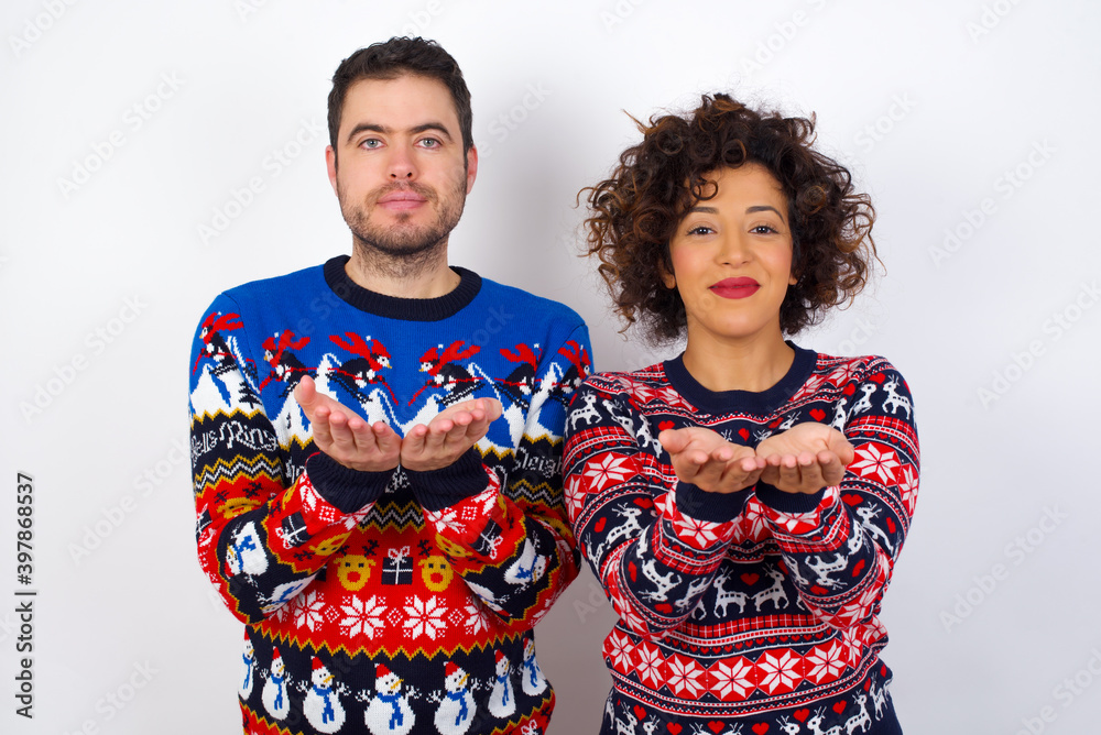 Young couple wearing Christmas sweater standing against white wall holding something with open palms, offering to the camera.