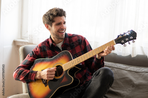 Young handsome artist playing the guitar and singing on the couch at home 