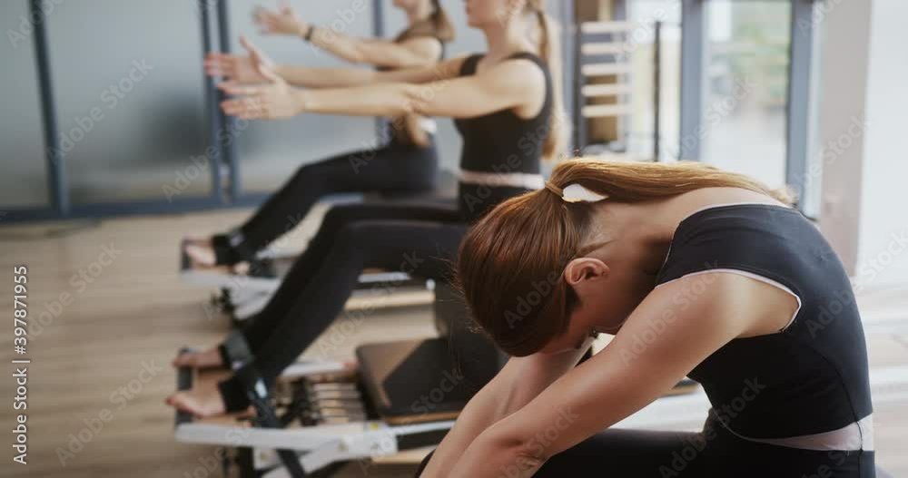 Woman stretching and resting during group pilates workout. Glad female bending forward and stretching back then breathing and turning head to camera against women doing pilates exercise during Woman stretching and resting during group pilates workout. Glad female bending forward and stretching back then breathing and turning head to camera against women doing pilates exercise during