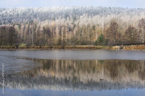Panorama of a forest with peaks covered with hoarfrost, partly illuminated by the sun with reflection in the water surface of a partially frozen lake, Brdy Protected Landscape Area (CHKO Brdy)