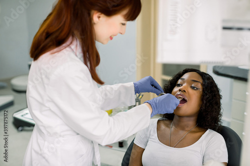 Young African female patient at dental office  opening mouth while smiling woman dentist in uniform and blue latex gloves  checking condition of teeth  using dental mirror. Oral care concept