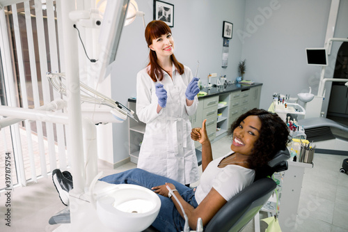 Healthy teeth and caries prevention concept. Young pretty African woman at the dentist s chair during a dental examination and treatment  showing thumb up. Young doctor dentist with dental tools