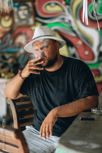 Man in full bloom  in a black and white panama hat and a bread on his face  sitting on a chair in a bar and smoking a cigar  filling the whole space with smoke. Portrait.