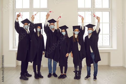 Group of successful diverse university graduates in face masks holding up their diplomas