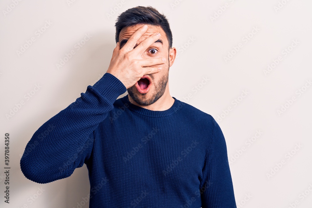 Young handsome man with beard wearing casual sweater standing over white background peeking in shock covering face and eyes with hand, looking through fingers afraid Young handsome man with beard wearing casual sweater standing over white background peeking in shock covering face and eyes with hand, looking through fingers afraid