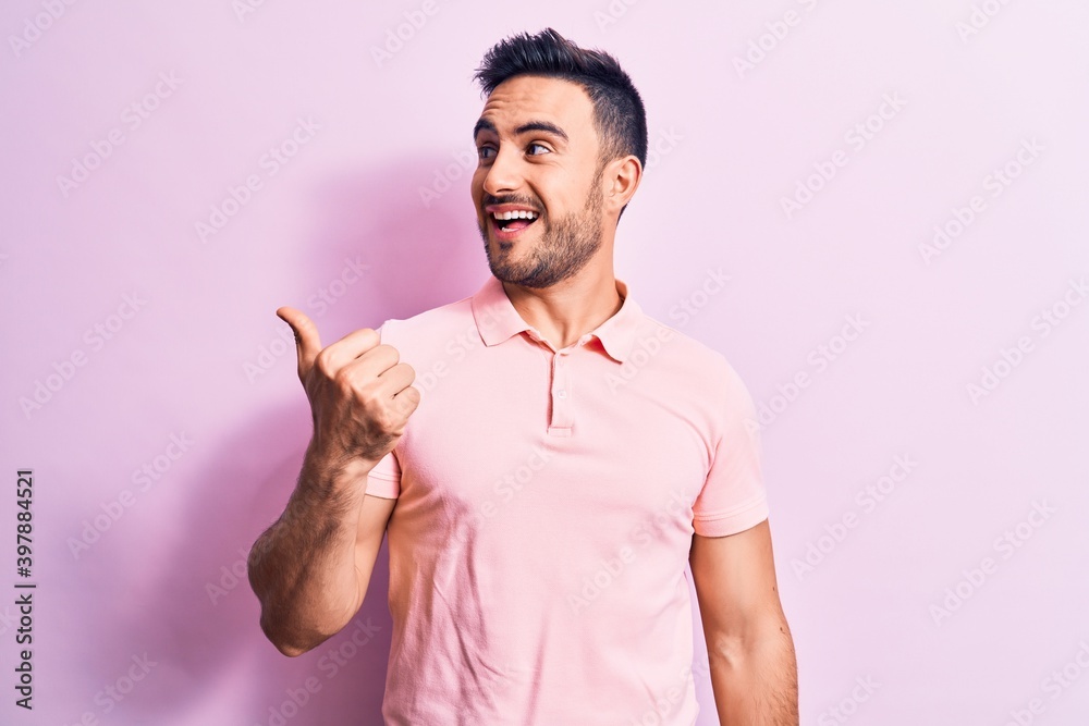 Young handsome man with beard wearing casual polo standing over pink background pointing thumb up to the side smiling happy with open mouth