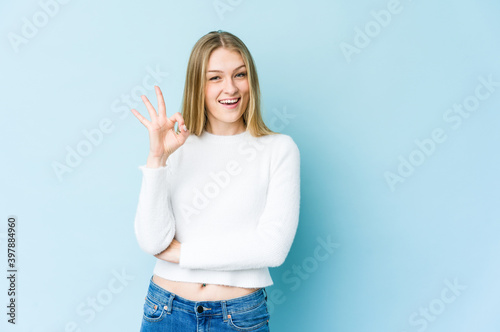 Young blonde woman isolated on blue background winks an eye and holds an okay gesture with hand.