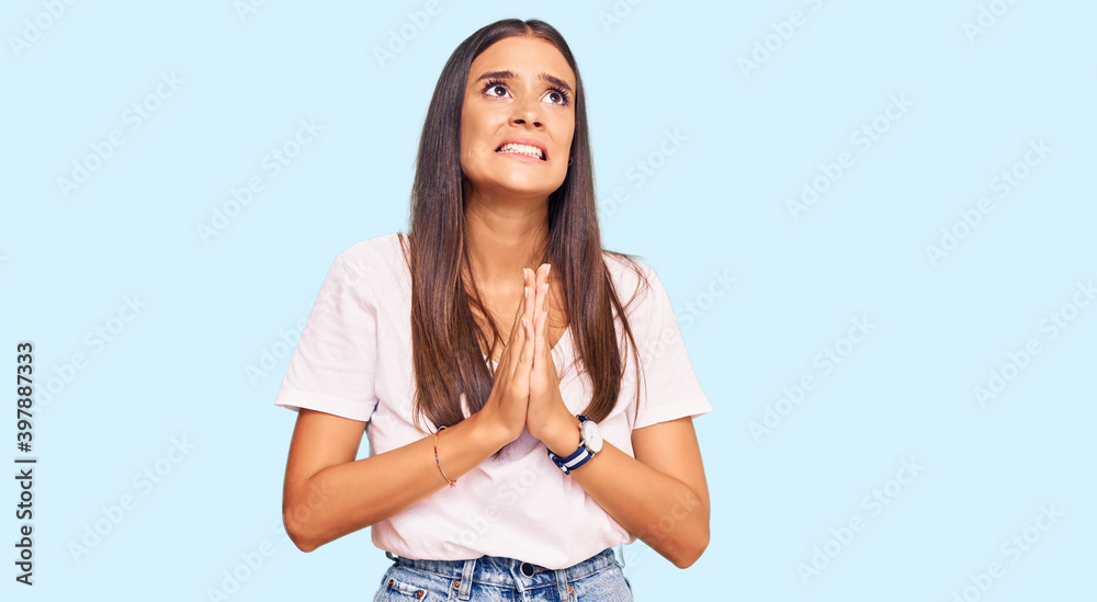 Young hispanic woman wearing casual white tshirt begging and praying with hands together with hope expression on face very emotional and worried. begging. Young hispanic woman wearing casual white tshirt begging and praying with hands together with hope expression on face very emotional and worried. begging.