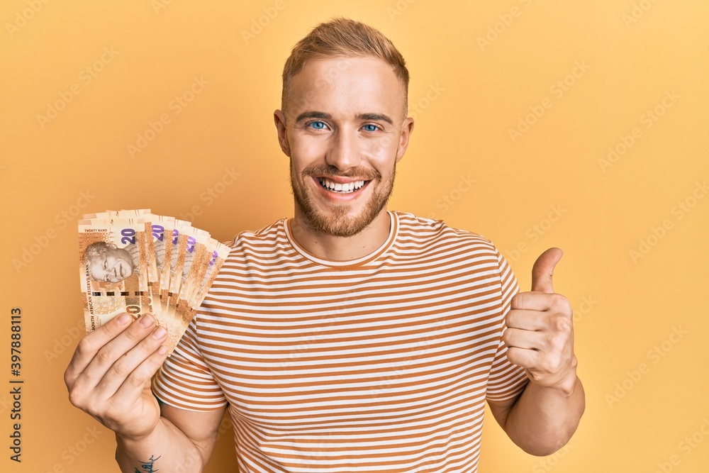 Young caucasian man holding south african 20 rand banknotes smiling happy and positive, thumb up doing excellent and approval sign Young caucasian man holding south african 20 rand banknotes smiling happy and positive, thumb up doing excellent and approval sign