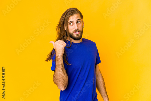 Young man with long hair look points with thumb finger away, laughing and carefree. © Asier