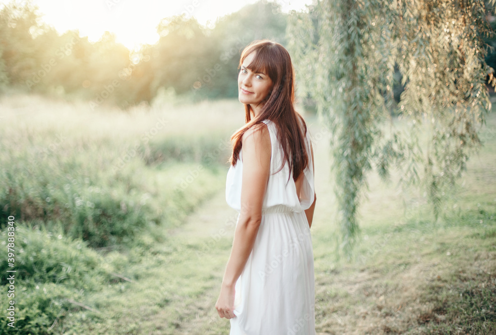 Beautiful happy smiling middle age young Caucasian woman with long dark brown red hair. Casual style real woman female in long white dress outdoors in park nature. Beautiful happy smiling middle age young Caucasian woman with long dark brown red hair. Casual style real woman female in long white dress outdoors in park nature.