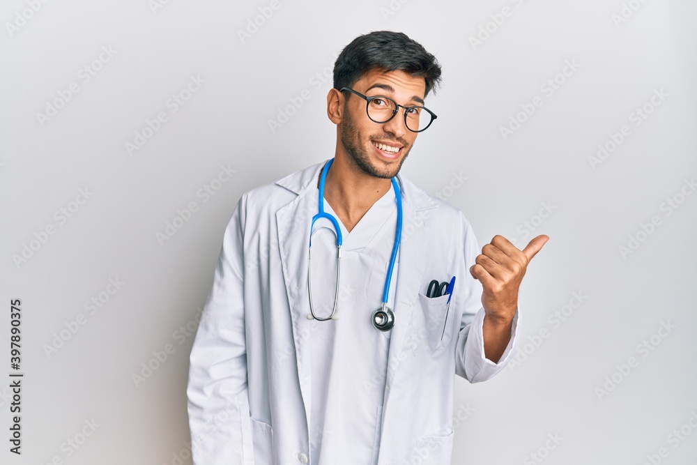 Young handsome man wearing doctor uniform and stethoscope smiling with happy face looking and pointing to the side with thumb up. Young handsome man wearing doctor uniform and stethoscope smiling with happy face looking and pointing to the side with thumb up.
