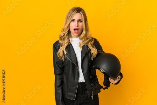 Young blonde caucasian biker woman holding helmet screaming very angry and aggressive.