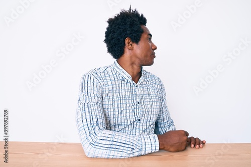 Handsome african american man with afro hair wearing casual clothes sitting on the table looking to side, relax profile pose with natural face with confident smile.