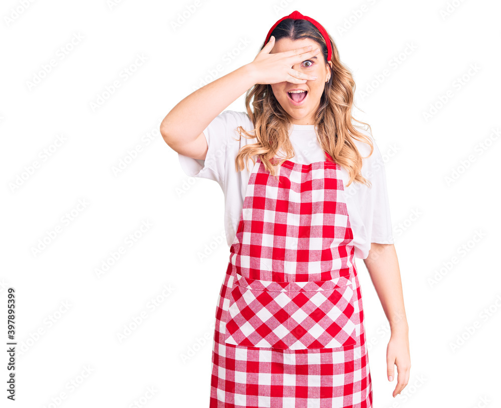 Young caucasian woman wearing apron peeking in shock covering face and eyes with hand, looking through fingers with embarrassed expression. Young caucasian woman wearing apron peeking in shock covering face and eyes with hand, looking through fingers with embarrassed expression.